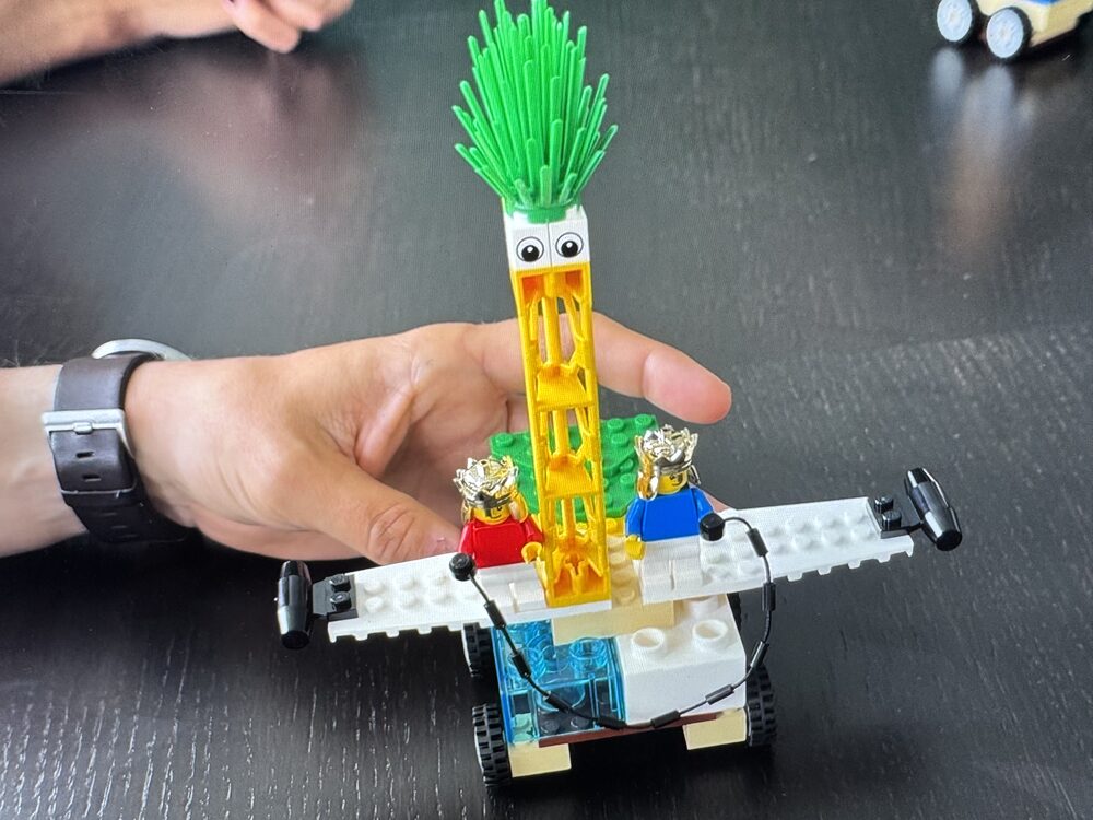 Modèle LEGO® SERIOUS PLAY® tenu dans une main : deux figurines couronnées sur une base à roues, dominées par une tour jaune coiffée d'yeux et de frondes vertes