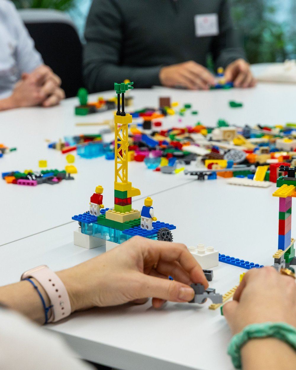 Mains de participants assemblant des briques autour d&rsquo;une table d&rsquo;atelier LEGO&reg; SERIOUS PLAY&reg;, une grue jaune et deux figurines pos&eacute;es sur un socle bleu au centre, briques en vrac &eacute;tal&eacute;es sur la table